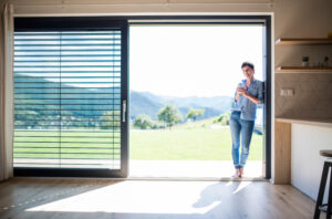 Front view of young woman with coffee standing by patio door at home.